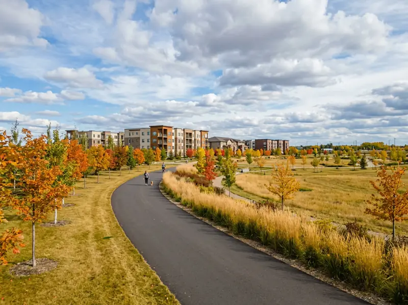 Paved pathway through an open park with young fall trees and low-rise condos behind in Blairmore