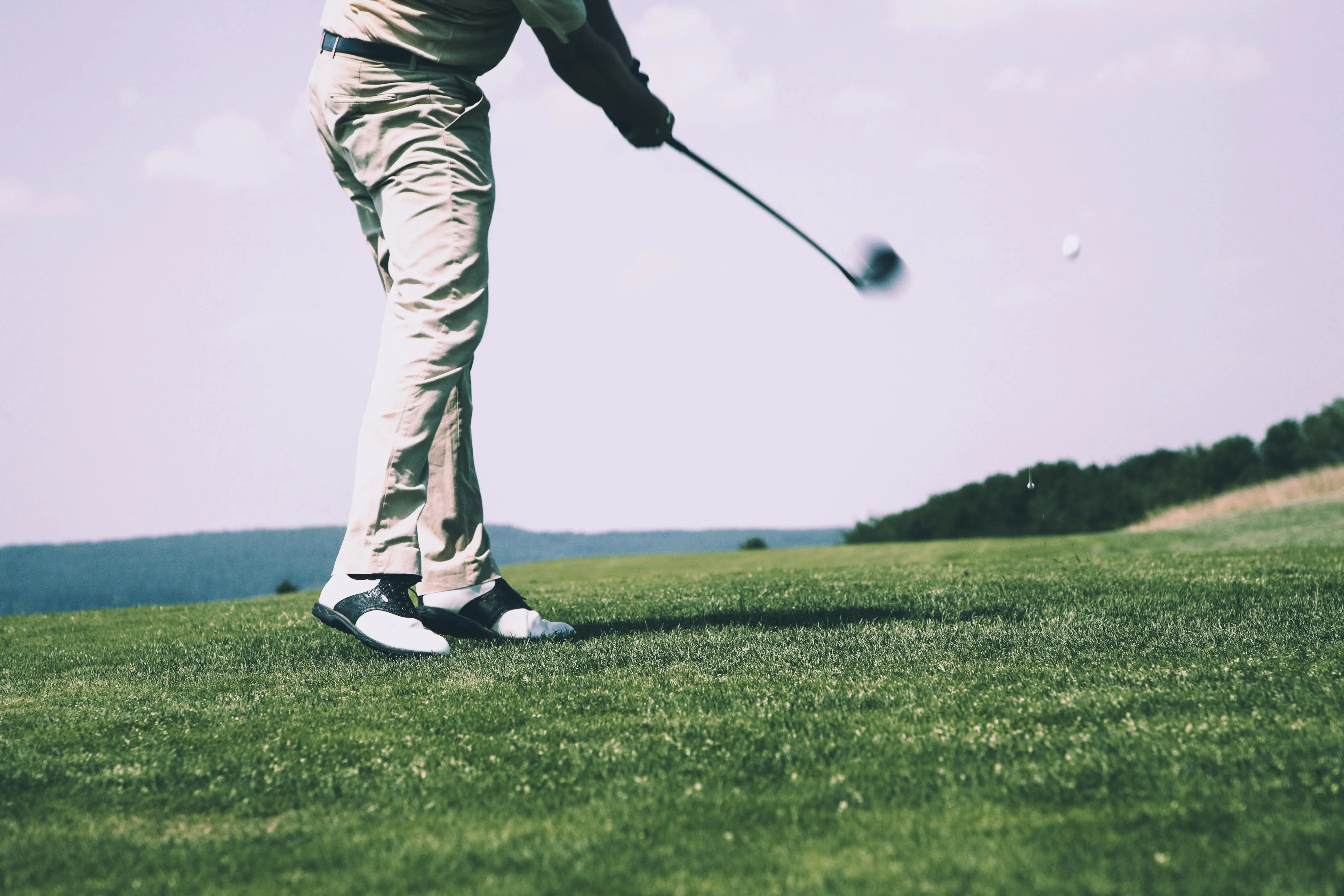 Golfer mid-swing on a sunny day, hitting a drive across a lush green fairway with forested hills in the background.