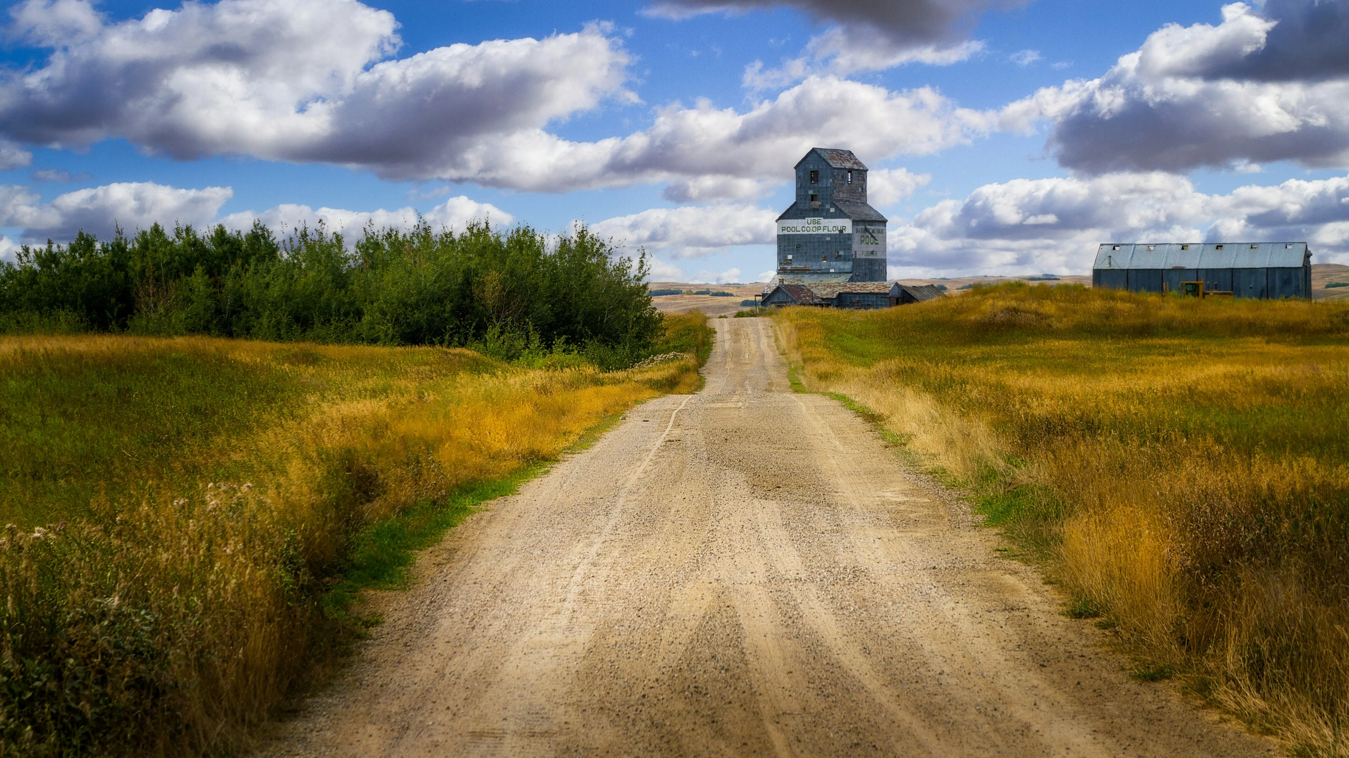 A rural dirt road leads toward an old wooden grain elevator under a partly cloudy blue sky, surrounded by golden and green prairie fields.